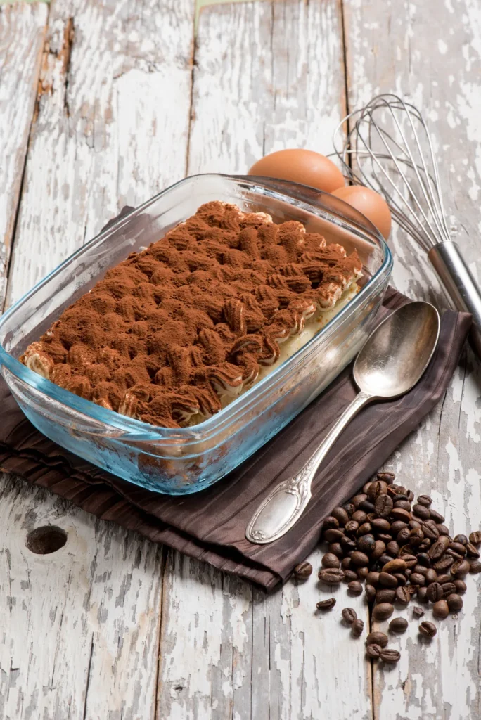 Ahşap masa üzerinde kahve çekirdekleri ve cam kapta kakao serpilmiş tiramisu
